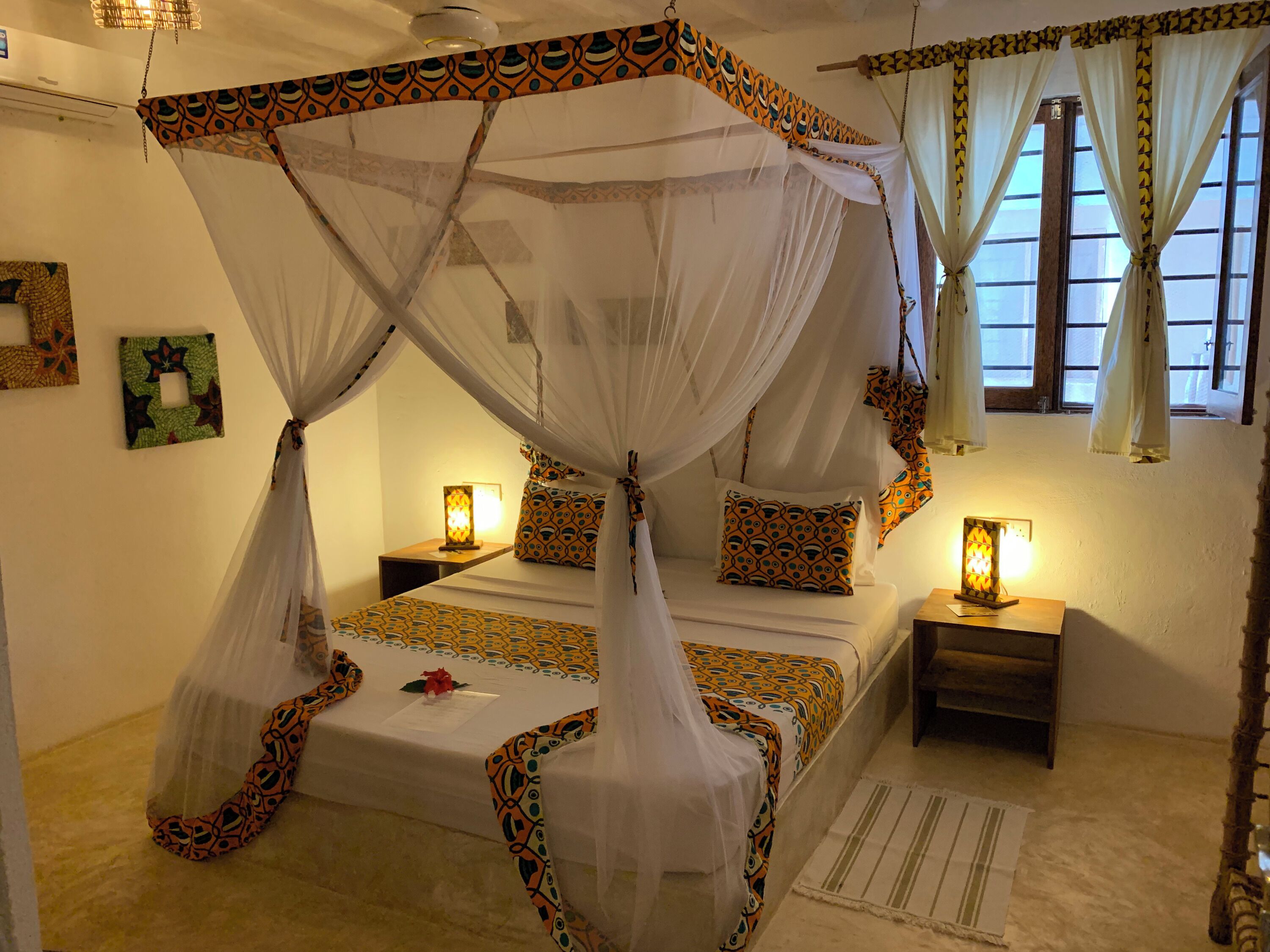 A room with a four-poster bed, two bedside tables with lamps, a decorative wall hanging, and a window with curtains.