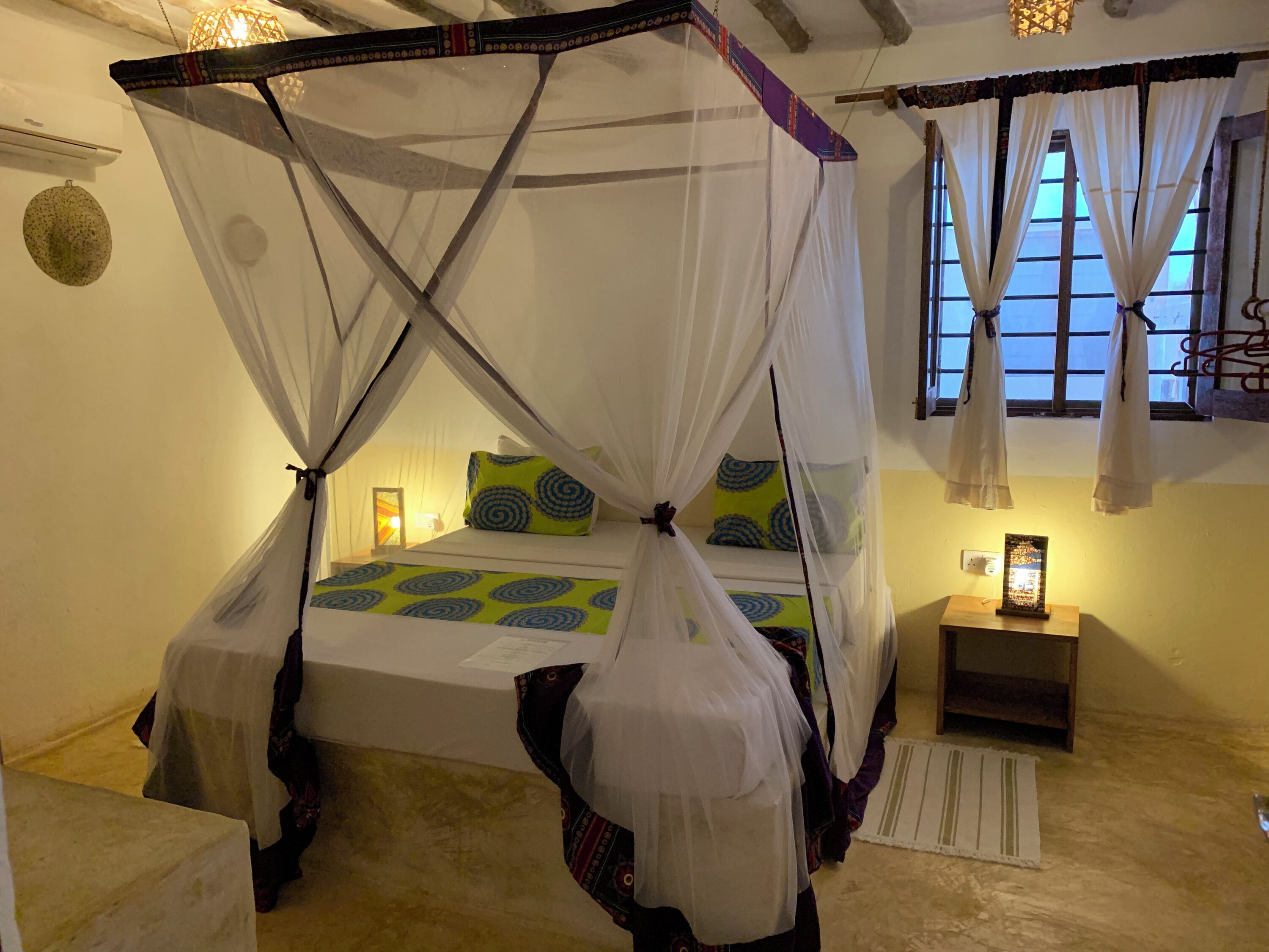 Ein Schlafzimmer mit einem Himmelbett, einem Nachttisch mit Lampe und einem Fenster mit Vorhängen.