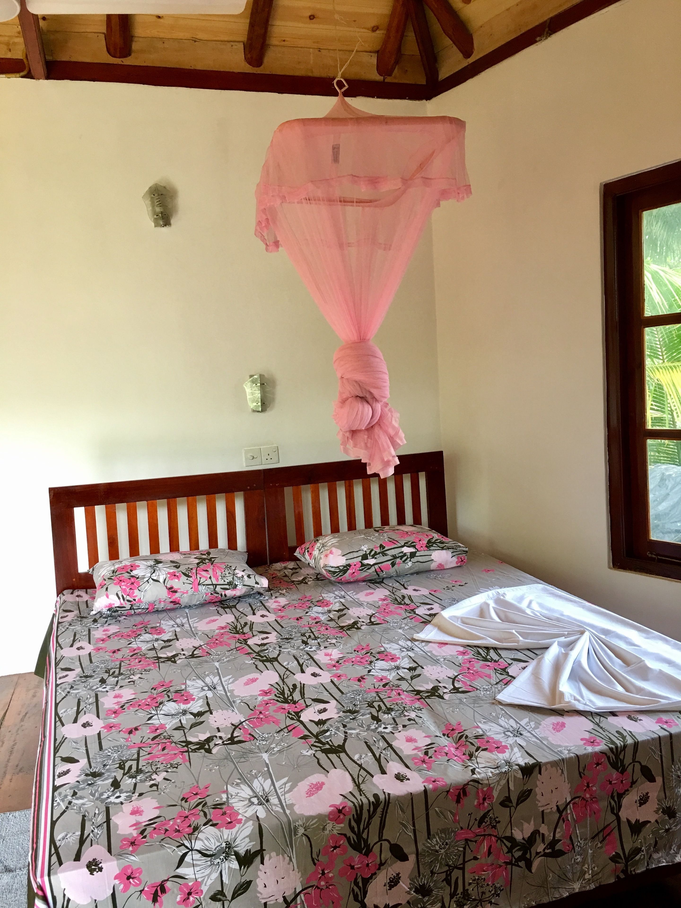 A bedroom with a wooden headboard, floral bedding, a mosquito net, and a window with a view of greenery.