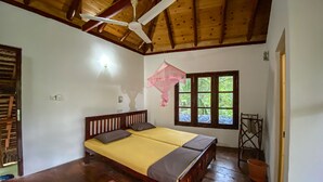 Una habitación con techo de madera, una cama con colcha amarilla, una ventana con vista a follaje y un ventilador de techo.