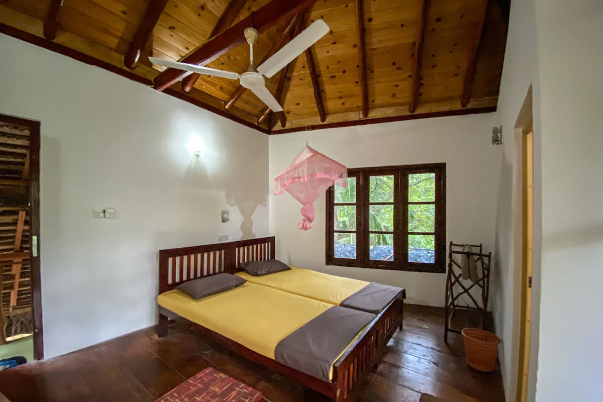 A room with a wooden ceiling, a bed with a yellow bedspread, a window with a view of greenery, and a ceiling fan.