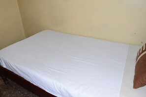 Un lit simple avec un matelas blanc et un oreiller marron, adossé à un mur uni.
