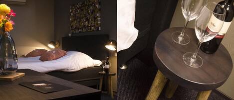 A room with a bed, a bedside table with a vase of flowers, a wine bottle, and glasses.