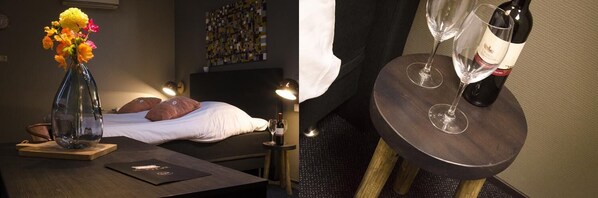 Una habitación con cama, mesita de noche con un florero, botella de vino y copas.