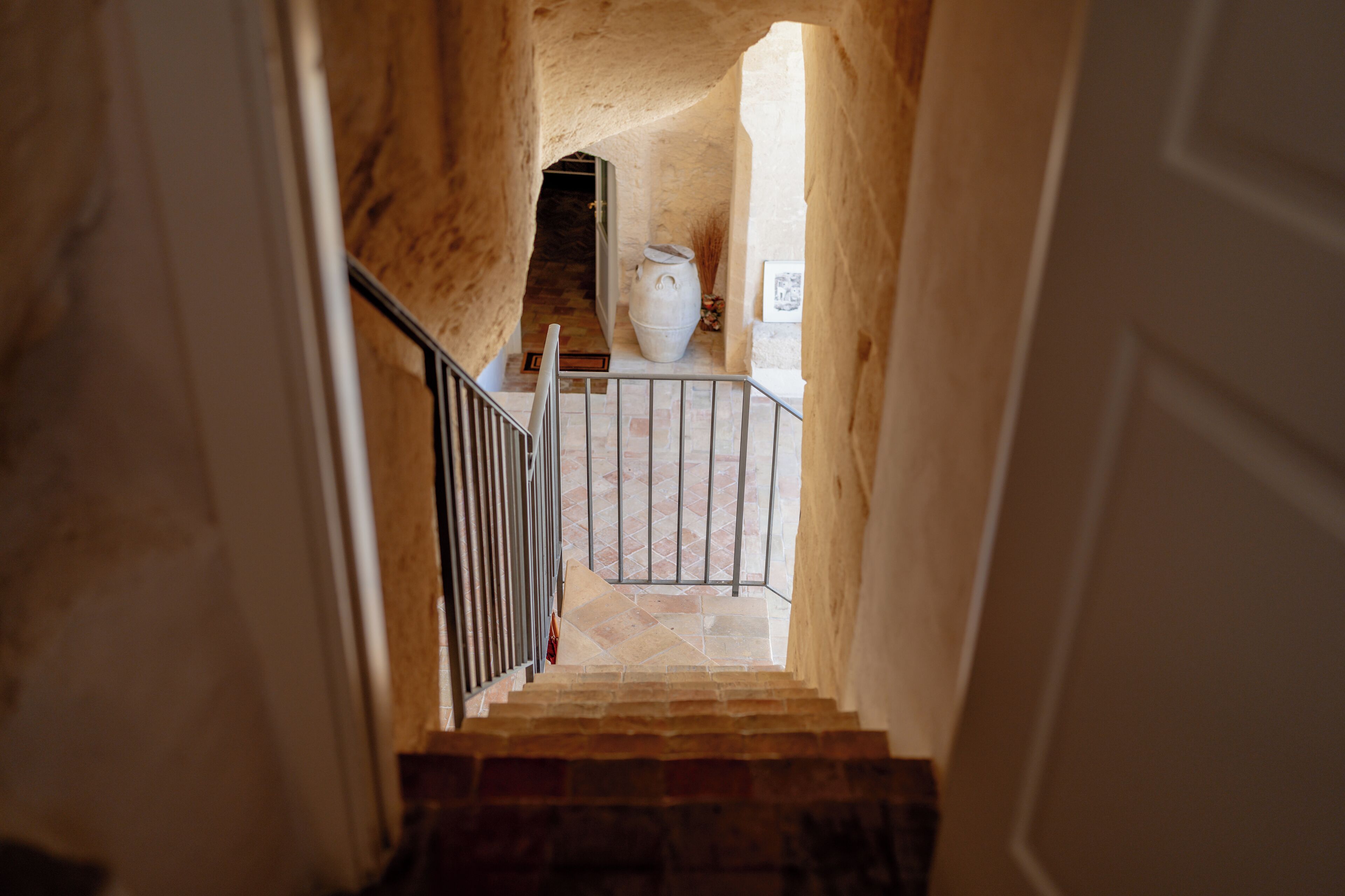 Un escalier menant à une pièce où se trouve un grand vase blanc, une rambarde en métal et un sol carrelé.