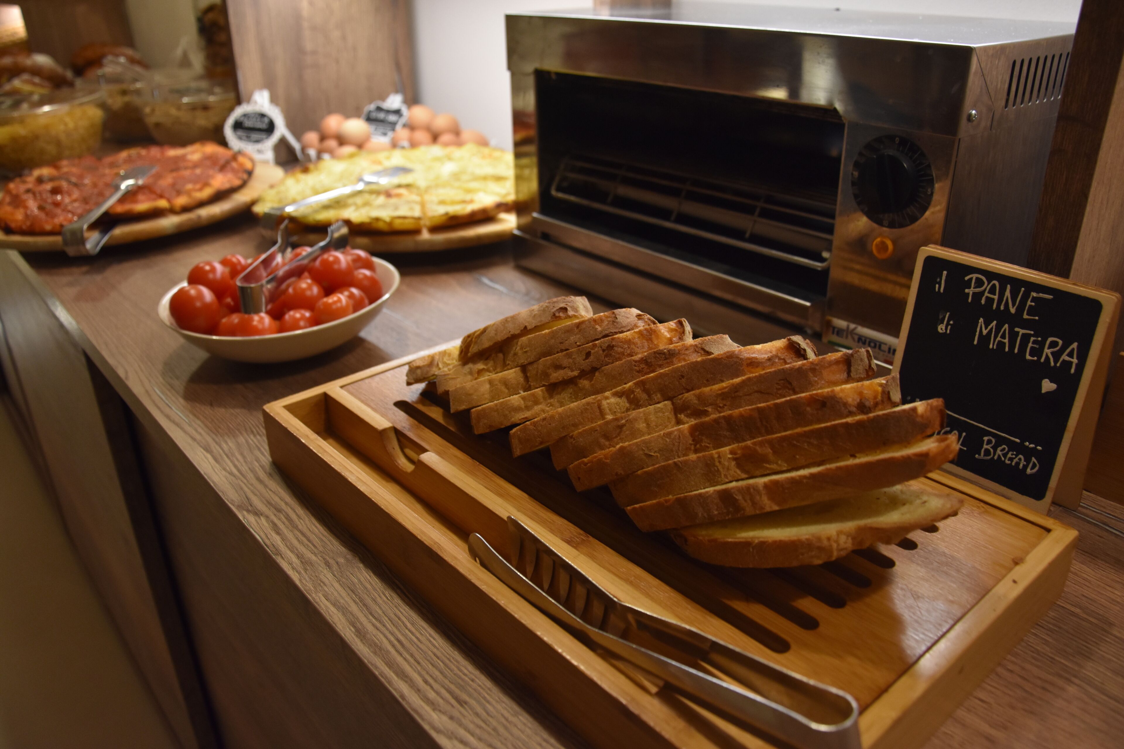 A buffet counter with various food items, including sliced bread, tomatoes, and a sign indicating 'Bread of Wheat'.