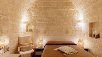 Quarto com paredes de pedra, cama com cobertor marrom, duas luminárias de cabeceira e uma cadeira com toalha branca.