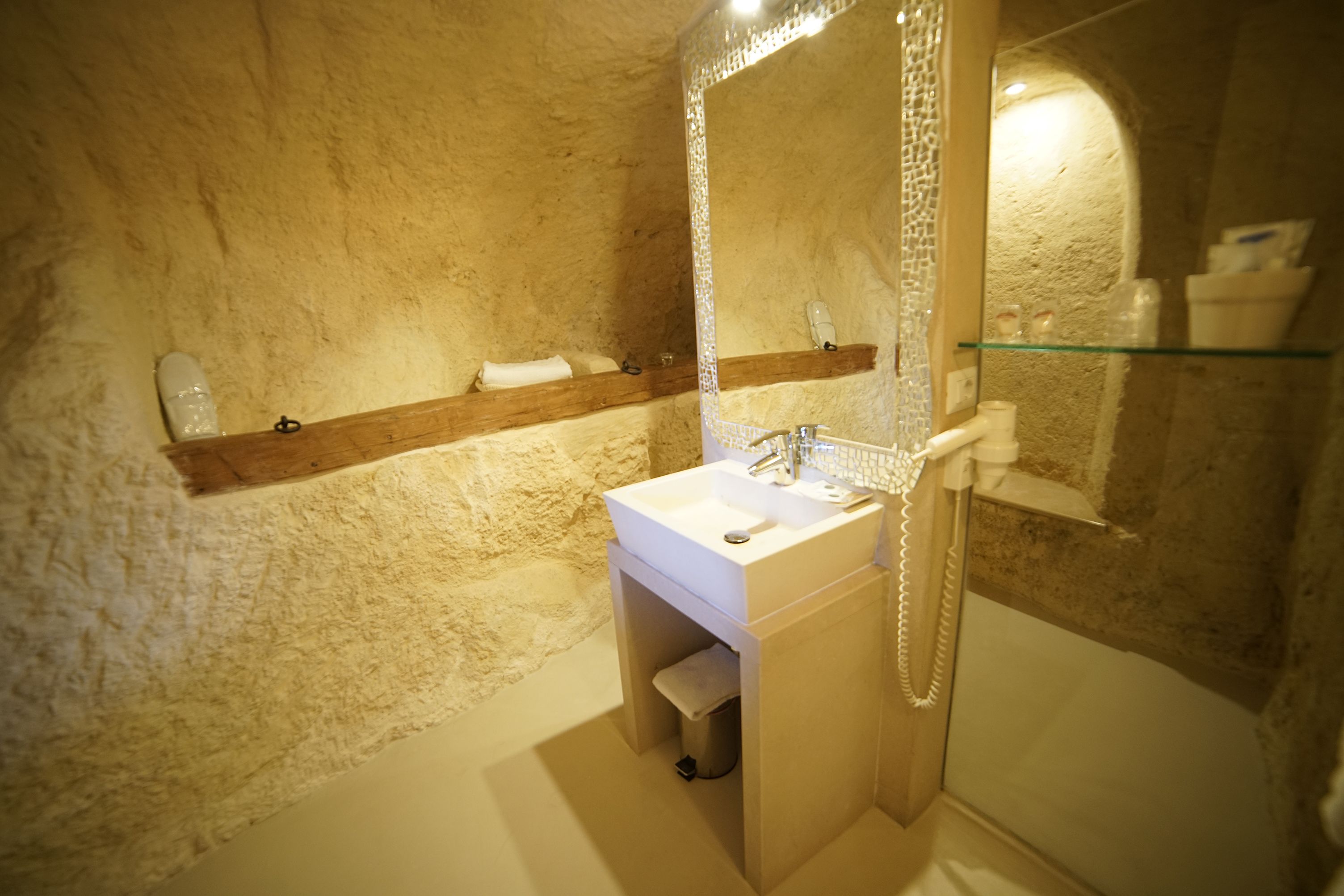Un baño con pared de piedra, estante de madera con toallas, lavamanos blanco y cabina de ducha de vidrio.