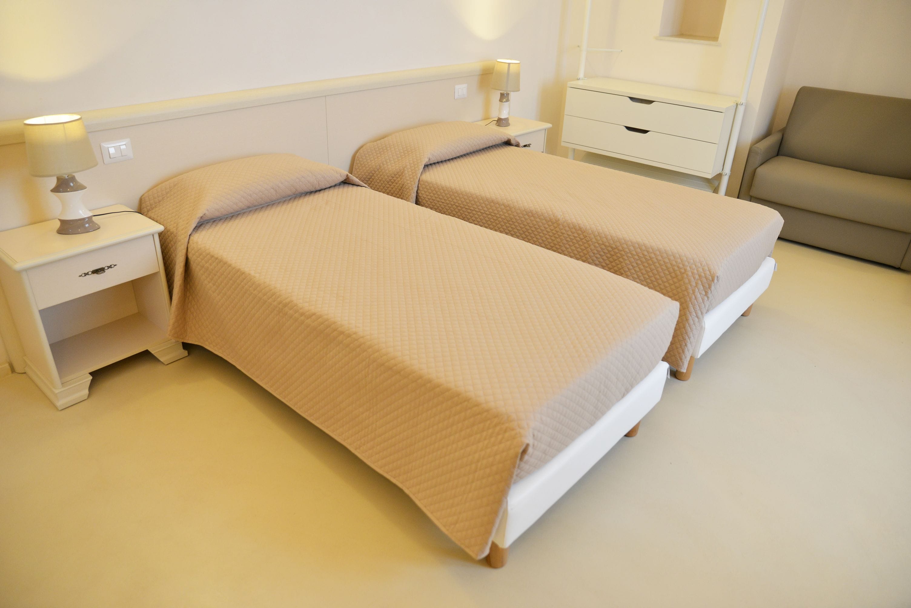 Habitación de hotel con dos camas individuales, una mesita de noche con lámpara, un cajón pequeño y un sofá.