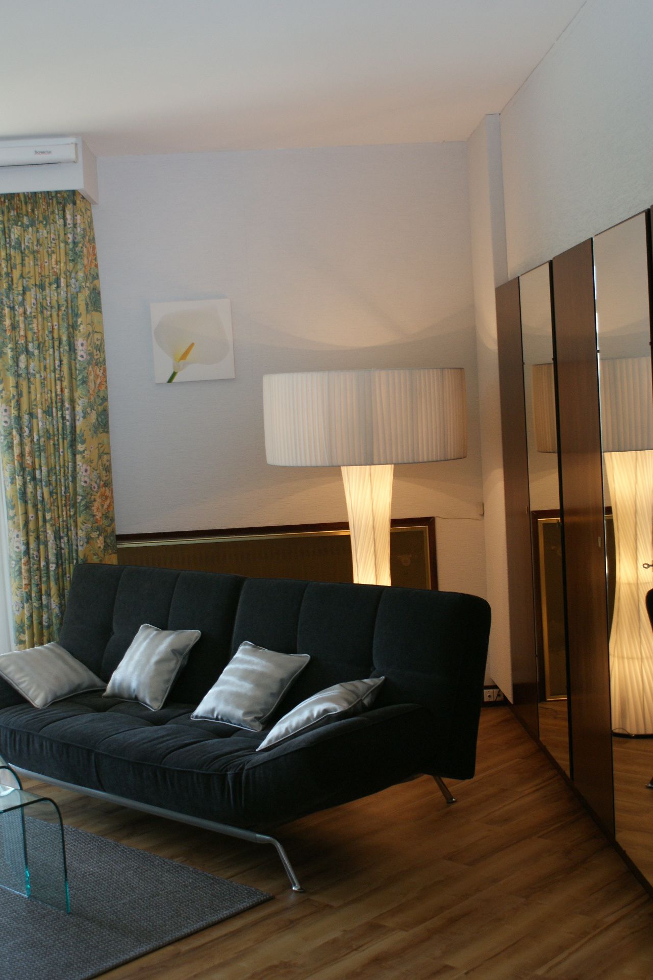A modern hotel room with a black sofa, a standing lamp, and a mirror cabinet.