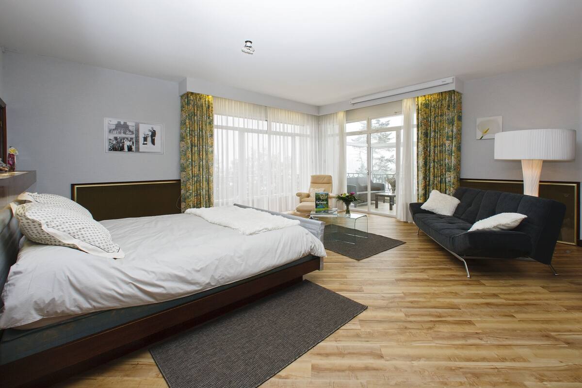 A modern hotel room with a large bed, a sofa, a dining table, and a view of the outdoors through large windows.
