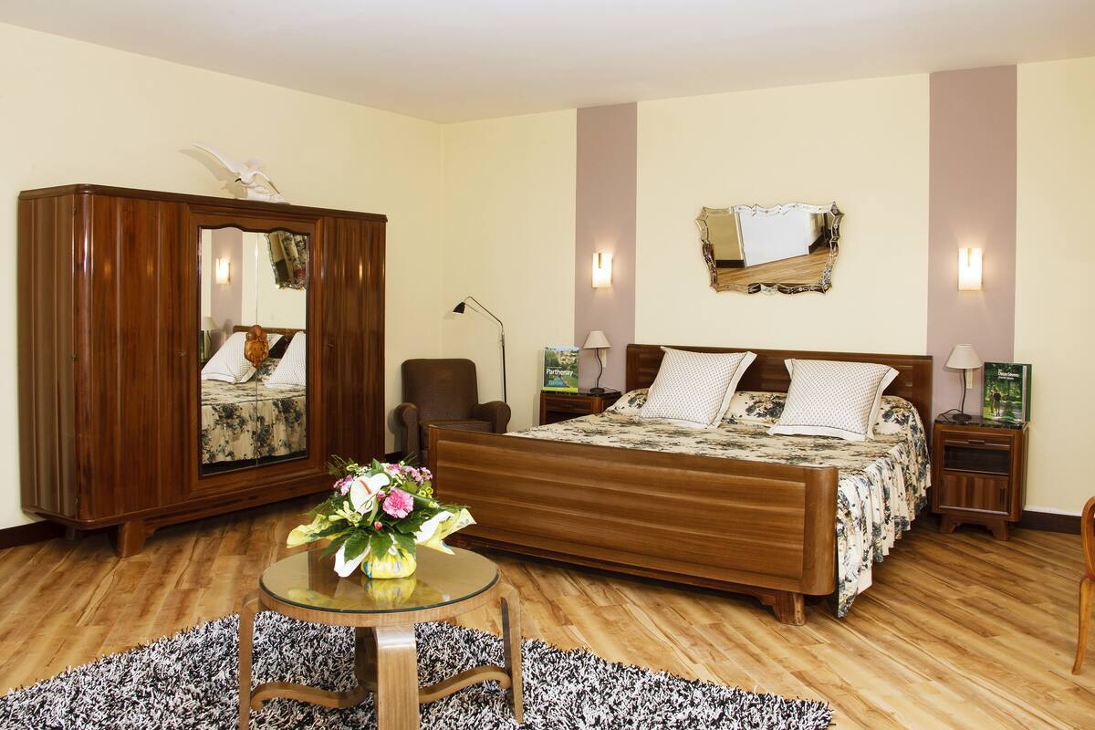 A bedroom with a wooden bed, a large mirror, a round glass table with a vase, and a small nightstand with a lamp.