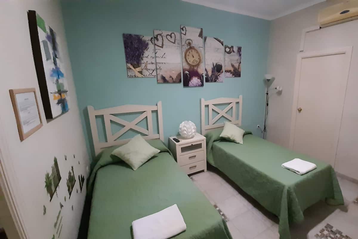 A bedroom with two beds, each with a green bedspread and white pillows. There is a white nightstand between the beds, and a wall clock above the nightstand. The room has light blue walls and a white door.