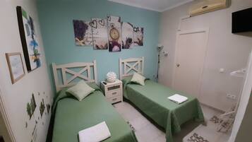 A bedroom with two beds, each with a green bedspread and white pillows. There is a white nightstand between the beds, and a wall clock above the nightstand. The room has light blue walls and a white door.