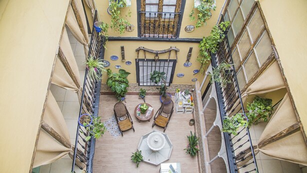 An indoor courtyard with a wooden floor, seating area with chairs and a table, potted plants, and a decorative window with a metal grille. - Pension Internacional (Córdoba)