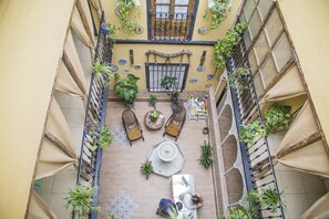An indoor courtyard with a wooden floor, seating area with chairs and a table, potted plants, and a decorative window with a metal grille. - Pension Internacional (Córdoba)