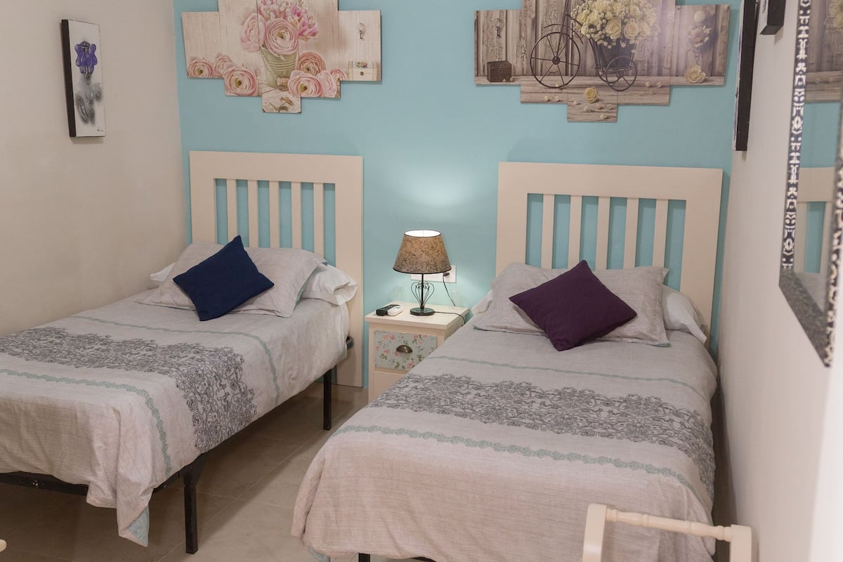 A bedroom with two beds, a nightstand with a lamp, and a small bench.