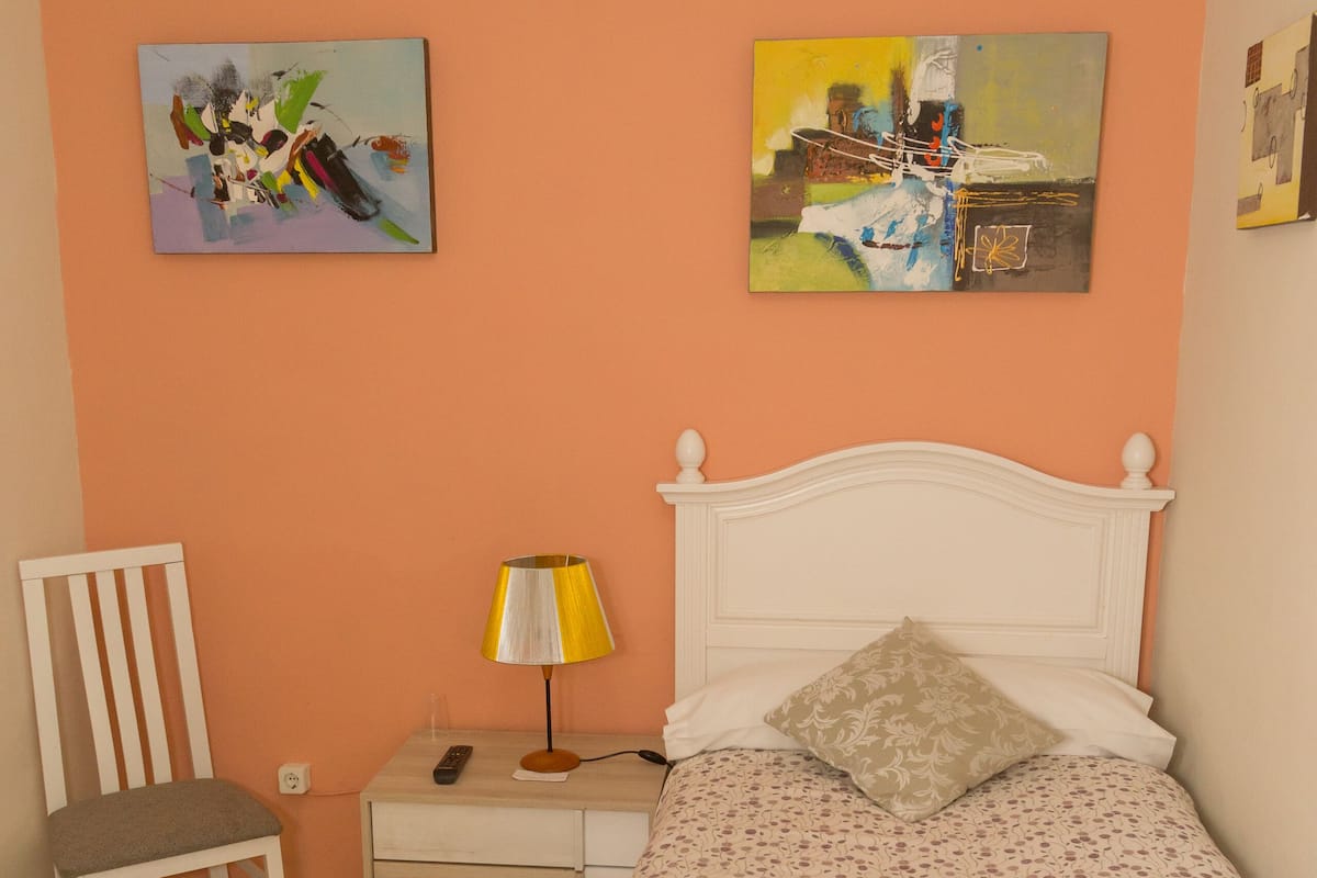 A bedroom with a bed, a chair, a nightstand, and two paintings on the wall.