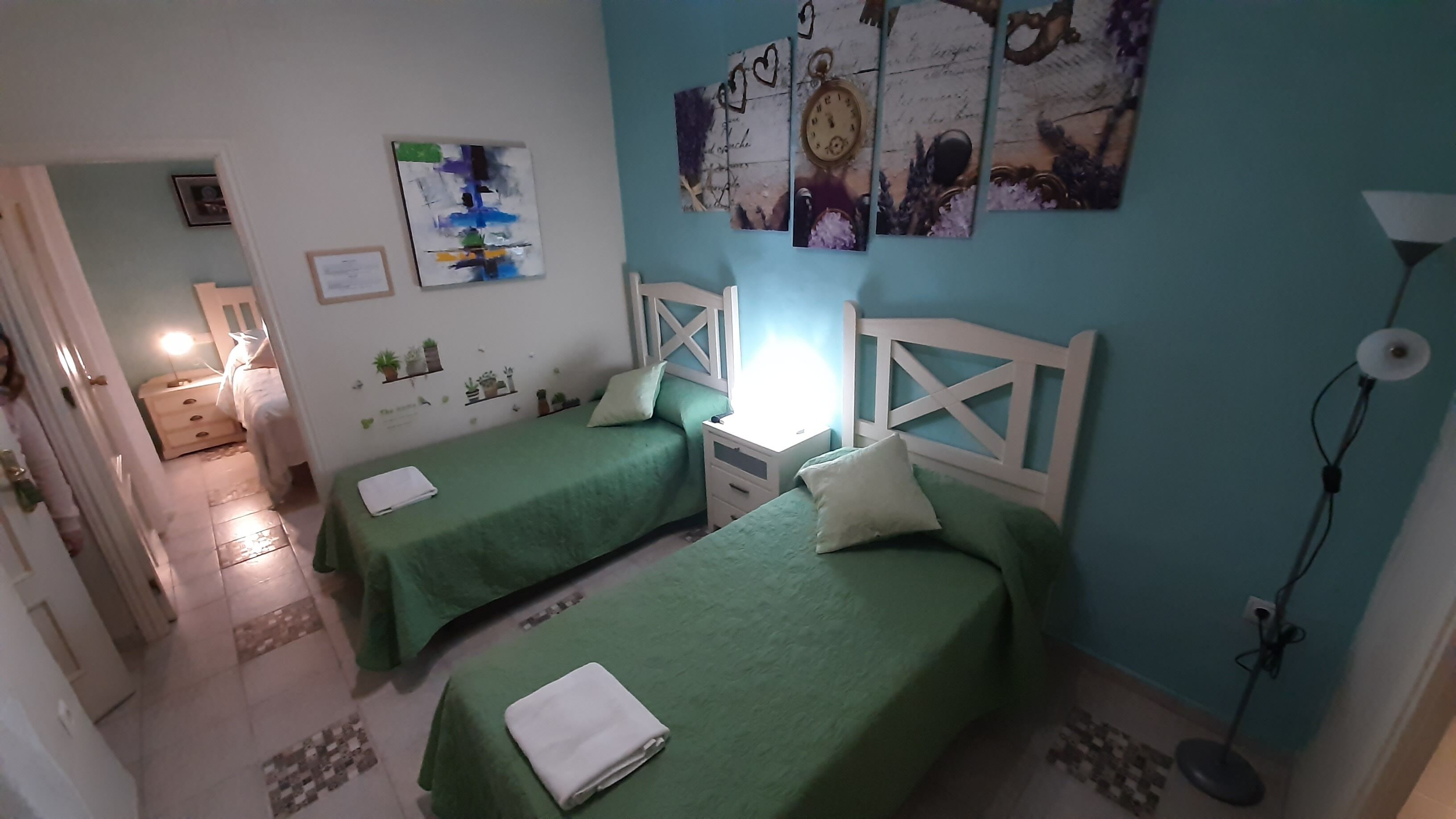 A room with two green beds, a white nightstand, a wall clock, and a small shelf with plants.