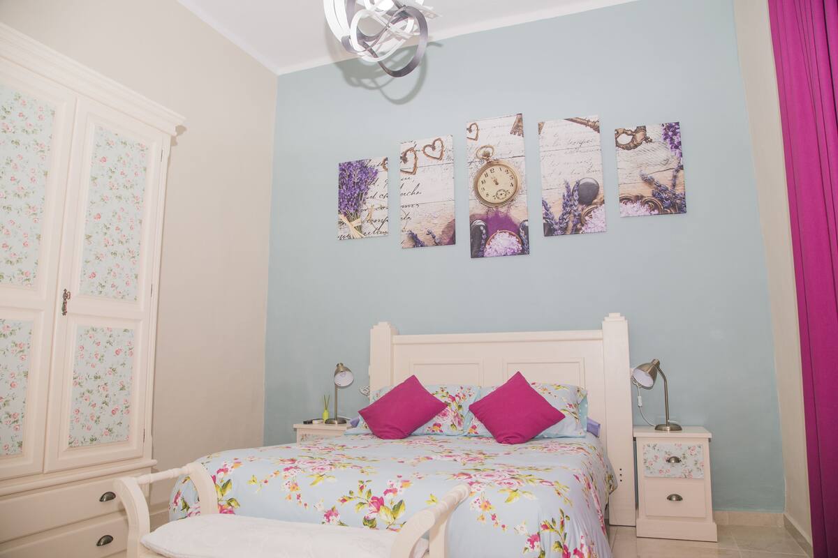 A bedroom with a floral bedspread, two purple pillows, a white headboard, a nightstand with a lamp, and a wall with three decorative pieces.