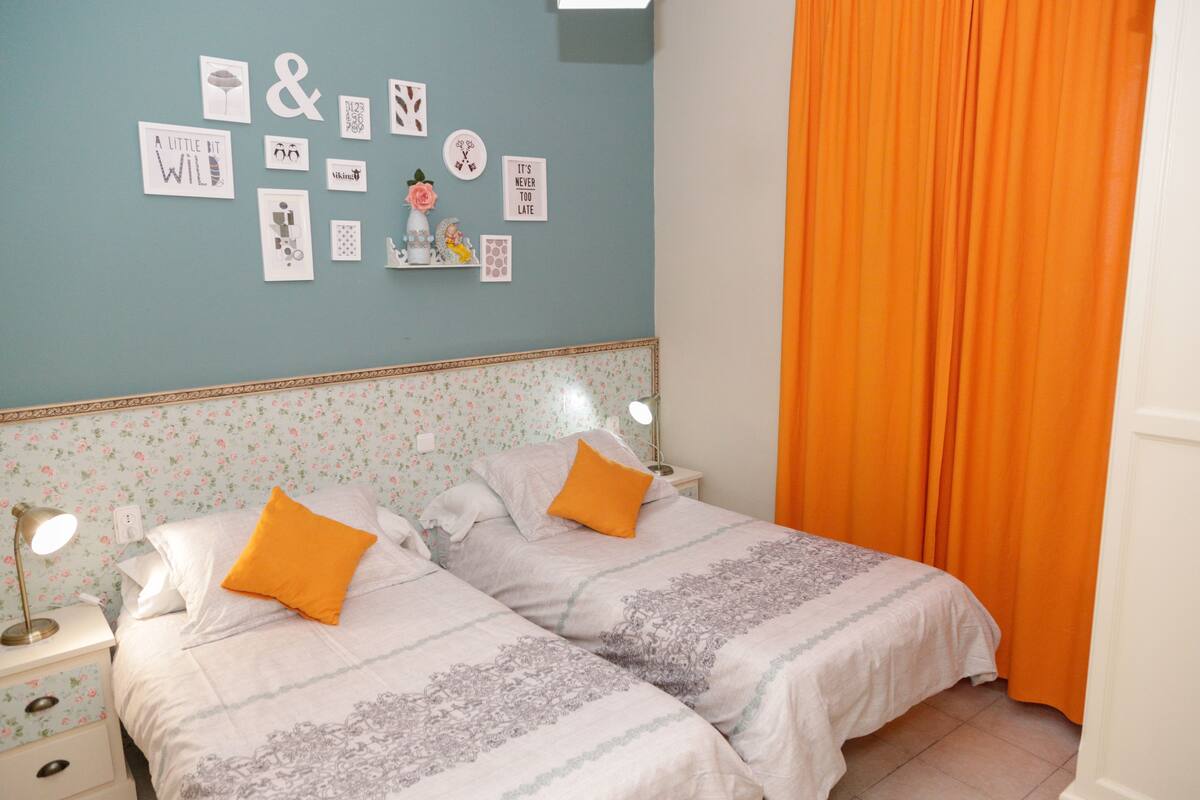 A bedroom with two beds, a floral wallpaper, a wall with various framed pictures, and an orange curtain.