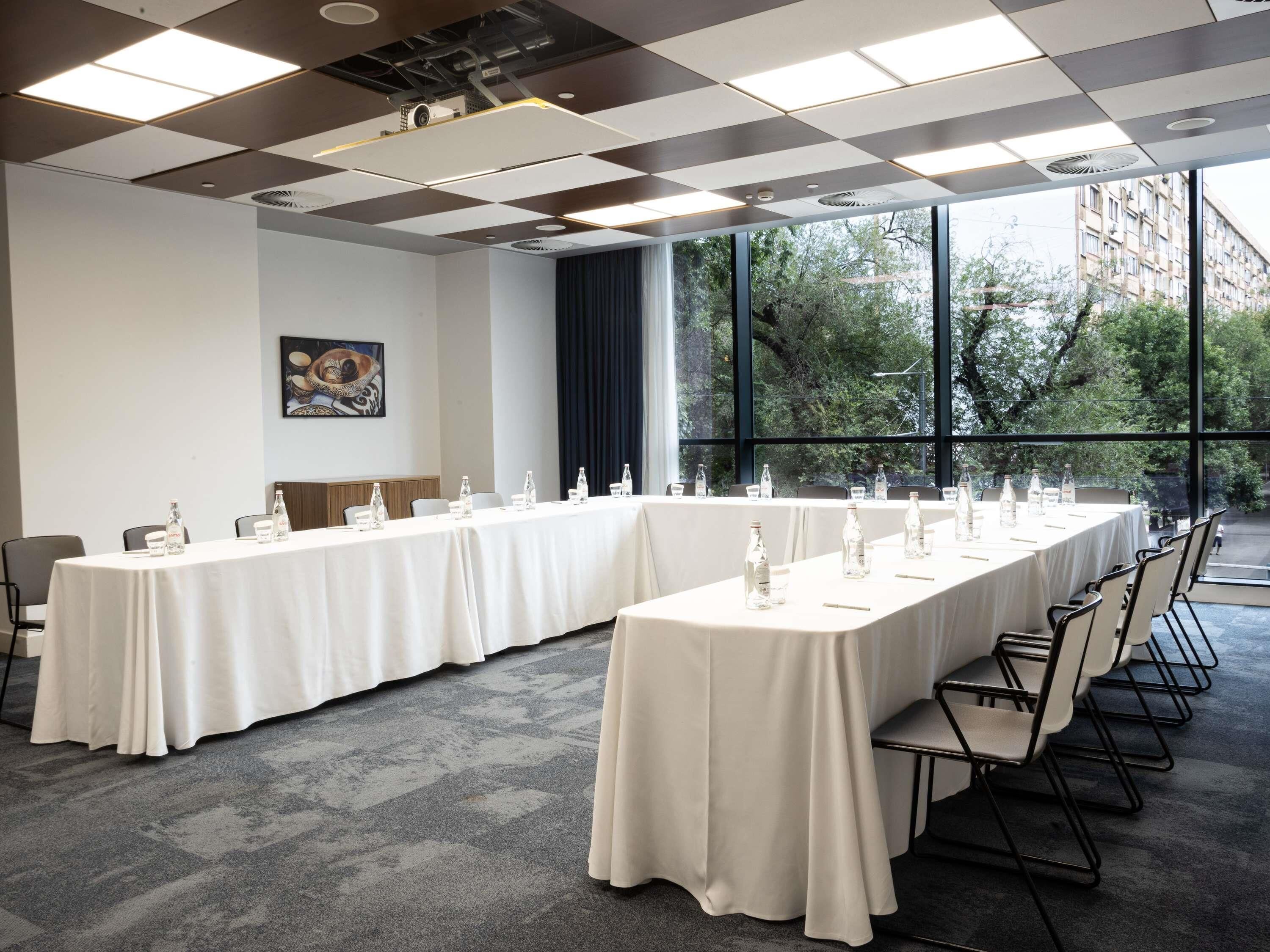 Une salle de conférence équipée de longues tables, de chaises et d’une grande fenêtre donnant sur des arbres et des immeubles.