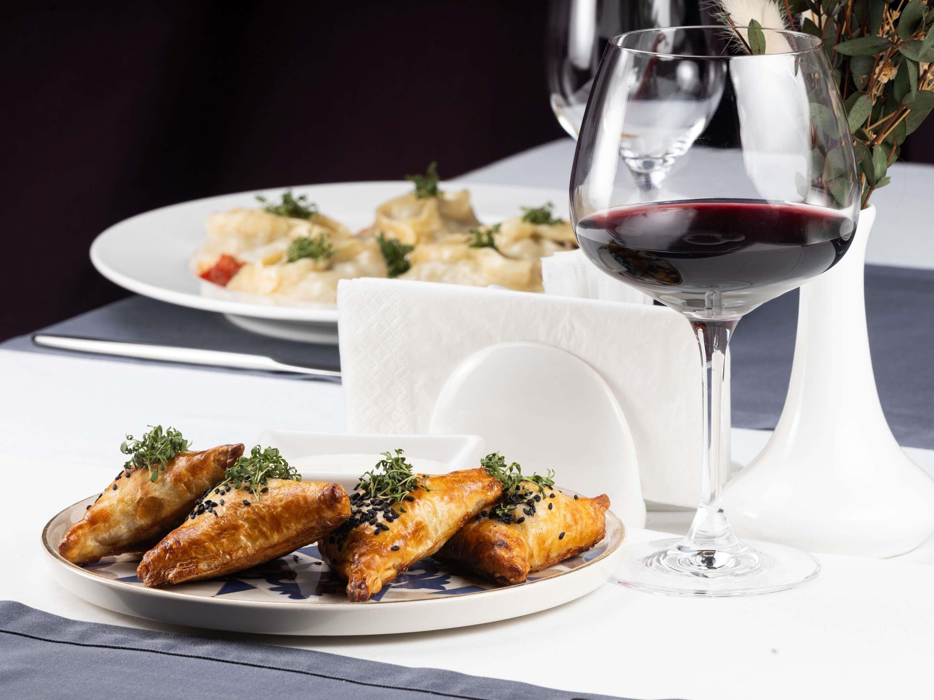 Una mesa de comedor con un plato de comida, una copa de vino tinto y un florero con follaje.
