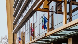 A modern building with a glass facade, multiple flags, and a covered entrance area. - Mercure Almaty City Center (Almaty)