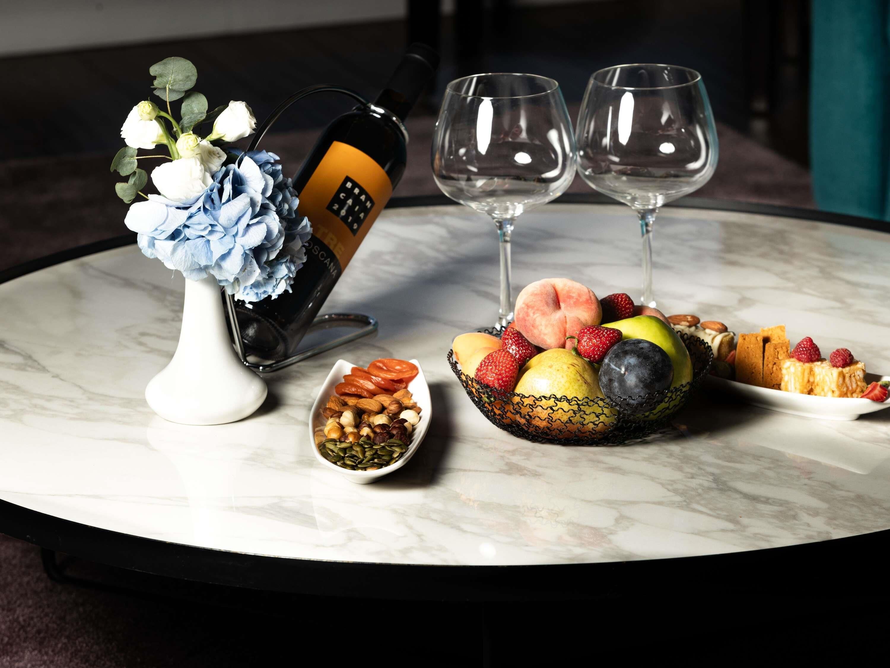 A marble table with a bottle of wine, two glasses, a bowl of nuts, a bowl of fruit, and a vase with flowers.