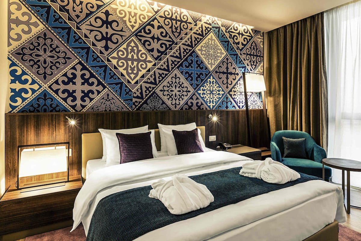 Una habitación de hotel con una cama grande, una mesita de noche, una lámpara, una silla y una pared con diseño.