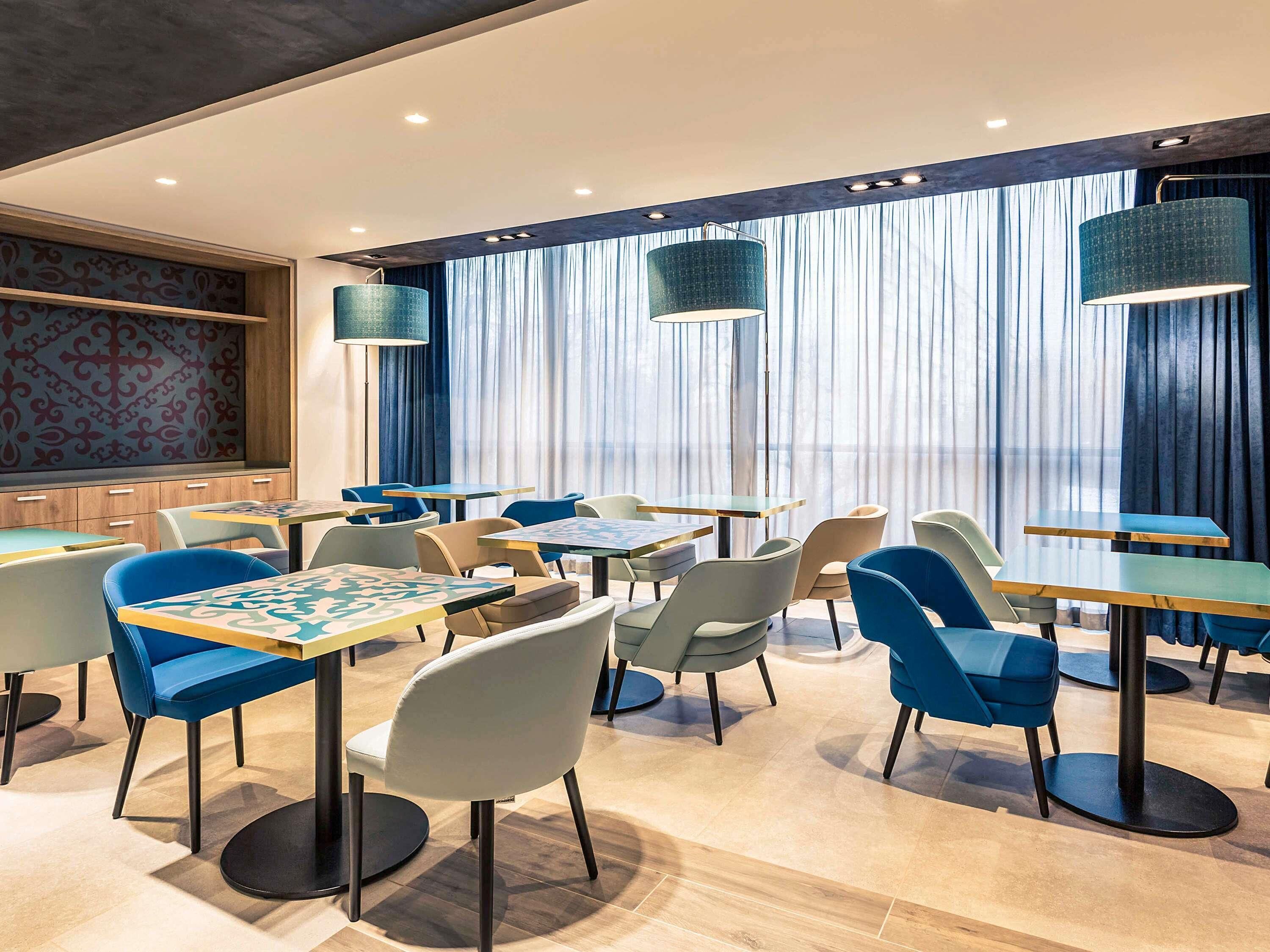 A modern dining area with tables and chairs, large windows with curtains, and pendant lighting.