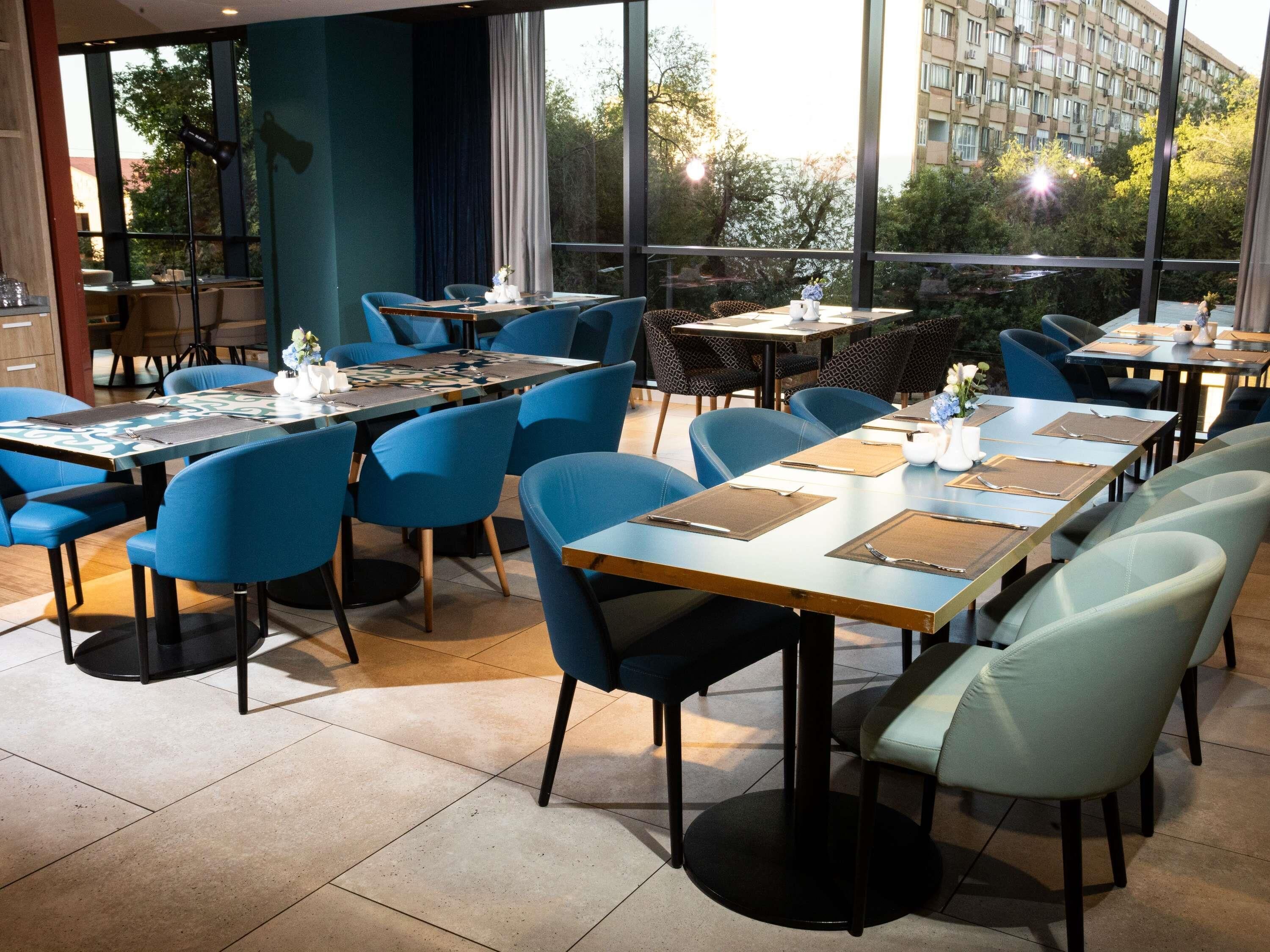 A restaurant with multiple tables and chairs, large windows, and a view of trees and buildings.