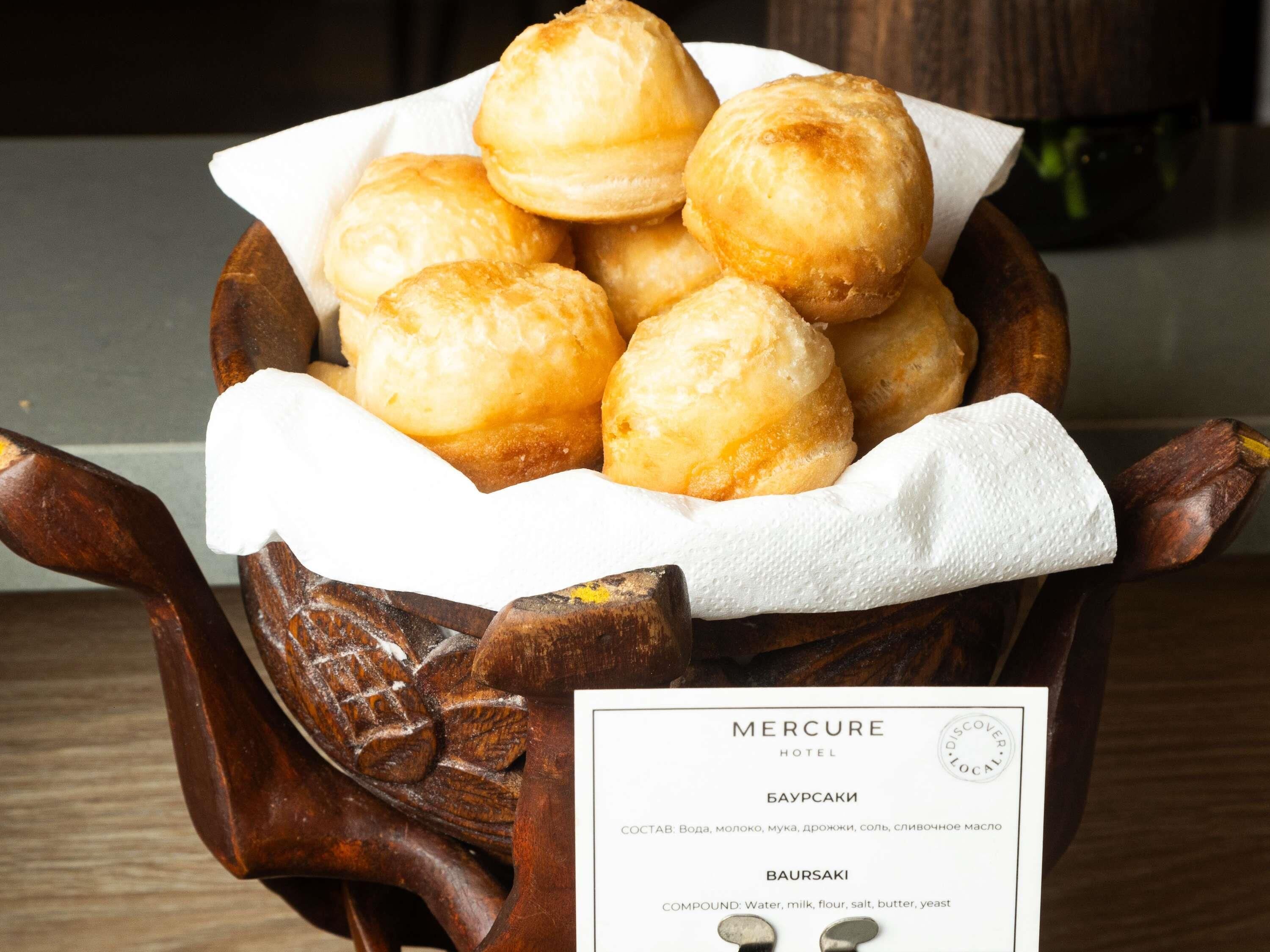 A wooden carved bowl filled with round pastries, labeled as 'Bauskiki' with a description of ingredients in a nearby sign.