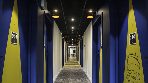 Een hotelgang met blauwe en gele wanden, genummerde deuren en een kamerbreed tapijt met dessin.