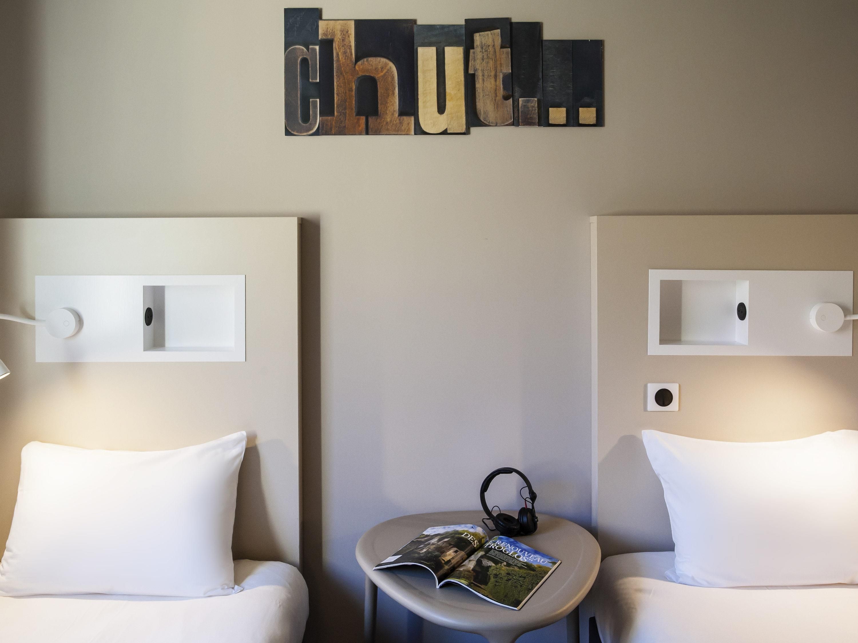 Habitación de hotel con dos camas, una mesita con una revista y auriculares, y una lámpara fijada a la pared.