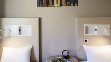Habitación de hotel con dos camas, una mesita con una revista y auriculares, y una lámpara fijada a la pared.
