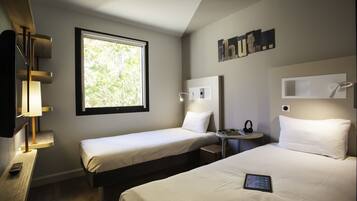 Habitación de hotel con dos camas, una mesita de noche con lámpara, una pequeña mesa con silla, una ventana que da a zonas verdes y un estante de pared con adornos.