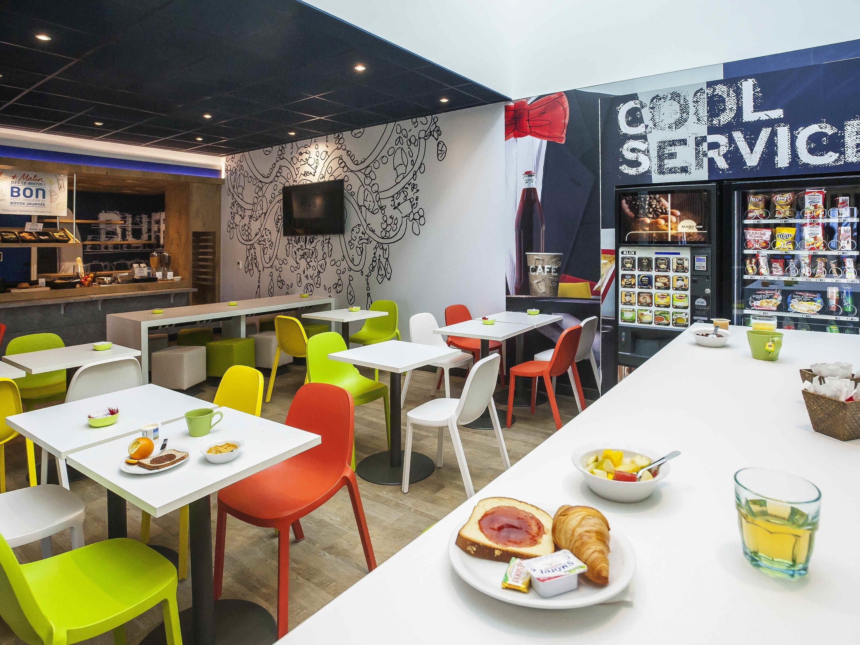 A café with tables and chairs, a vending machine, and a wall with a 'Good Service' sign.
