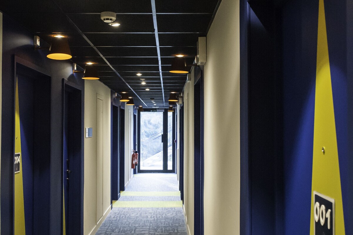 Un couloir d’hôtel avec un tapis à motifs, des murs bleus et jaunes, et un éclairage au plafond.