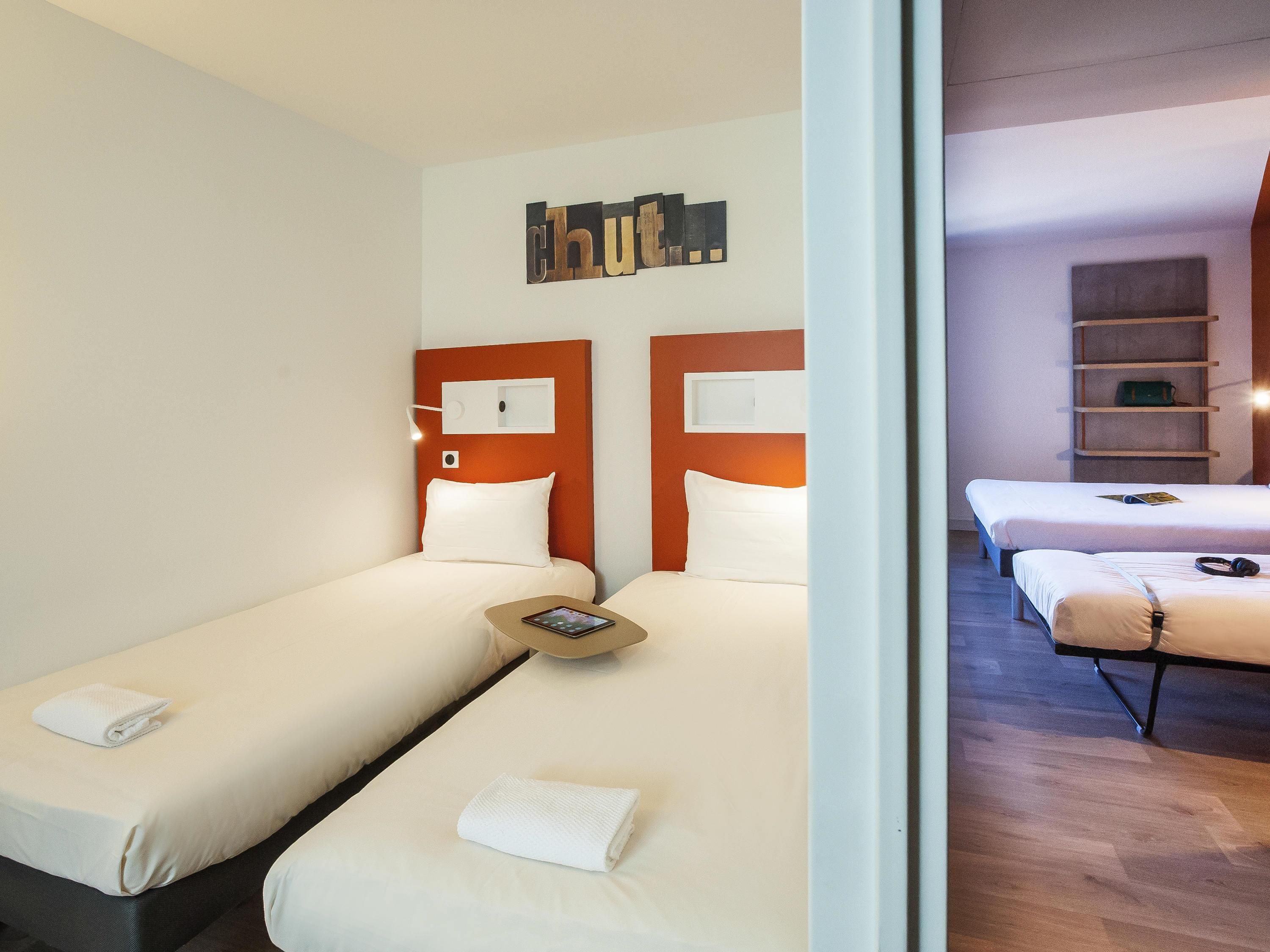A hotel room with two beds, a headboard with a clock, a small table, and a wall-mounted artwork.