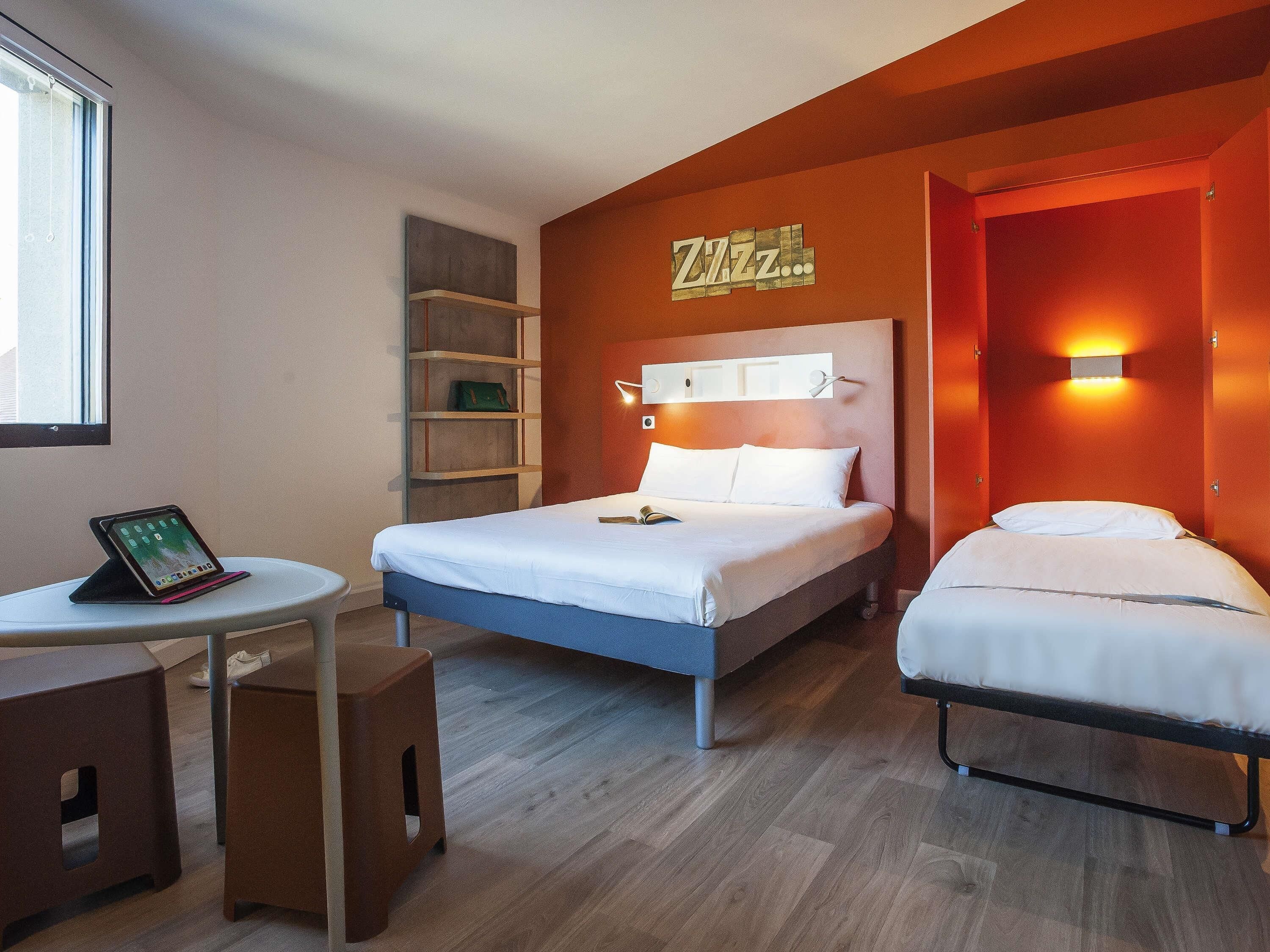 Habitación de hotel con cama, mesitas de noche, un escritorio con una tableta y una estantería en la pared.