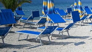 Beach chairs and umbrellas arranged on a pebbled beach with a clear view of the sea and distant hills. - Hotel Medena (Seget)