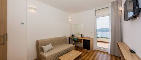 A modern room with a wooden floor, a beige quilted sofa, a wooden coffee table, a desk with a chair, a TV mounted on the wall, and a large window with a view.