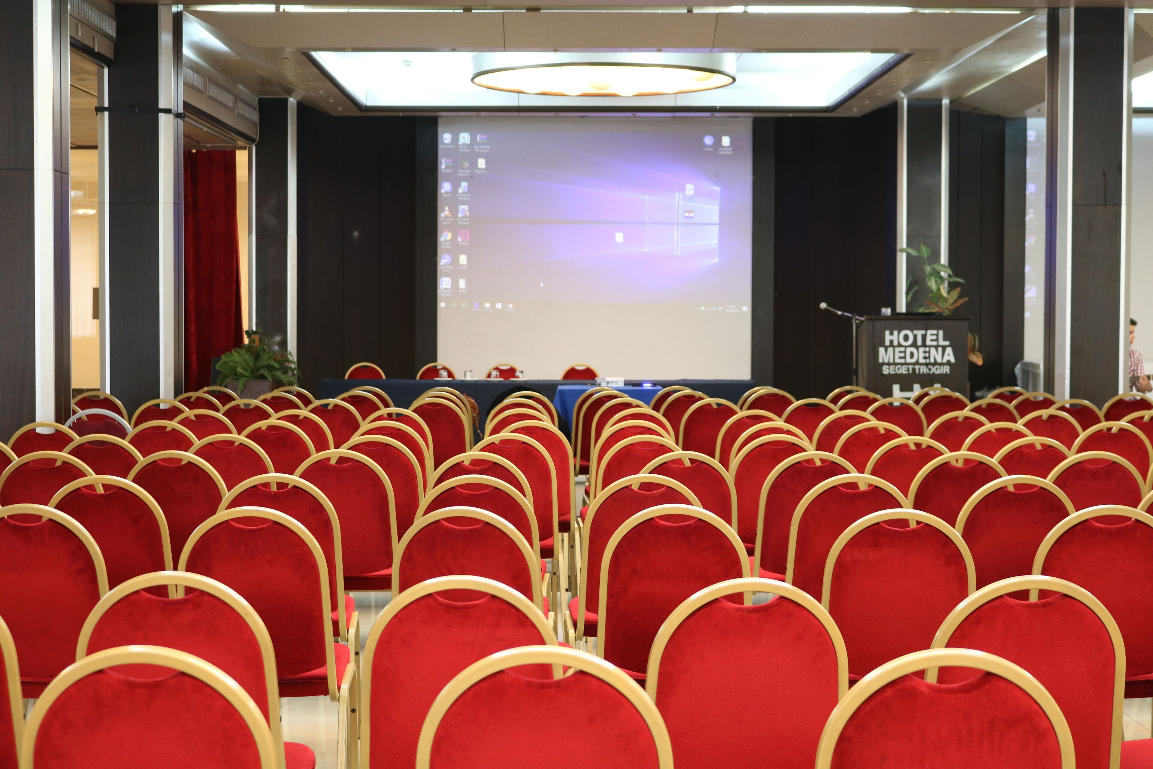 Ett konferensrum med rader av röda stolar, en talarstol med skylten ”HOTEL MEDENA” och en stor skärm som visar en presentation.