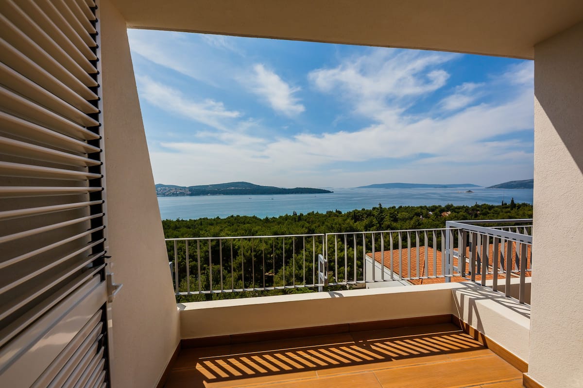 A balcony with a view of the sea and distant hills, wooden flooring, and a white railing.