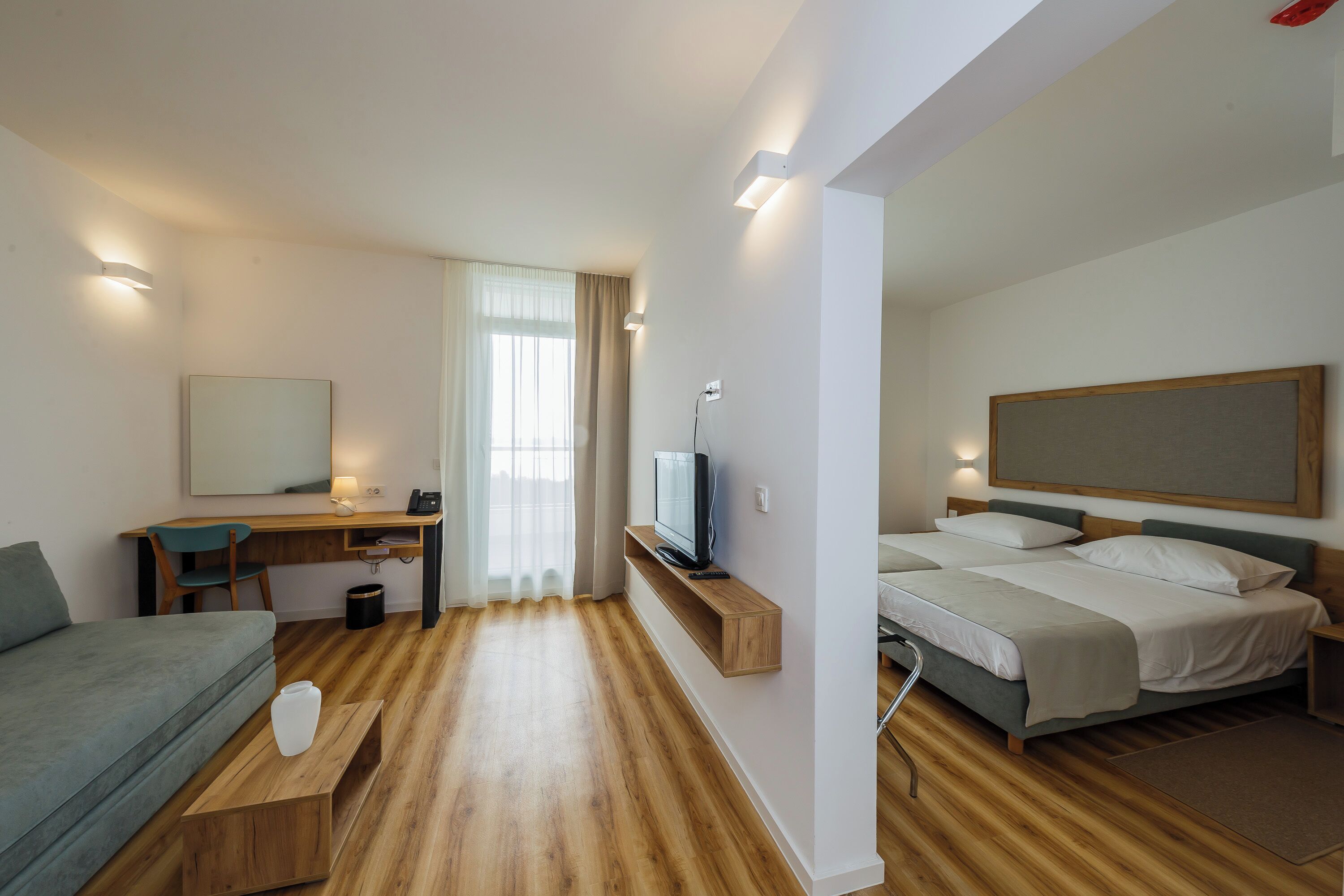 A modern hotel room with a wooden floor, a bed with white bedding, a desk with a television, and a large window with curtains.