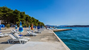 A beachside area with lounge chairs and umbrellas, a concrete walkway, and a calm sea. - Hotel Medena (Seget)