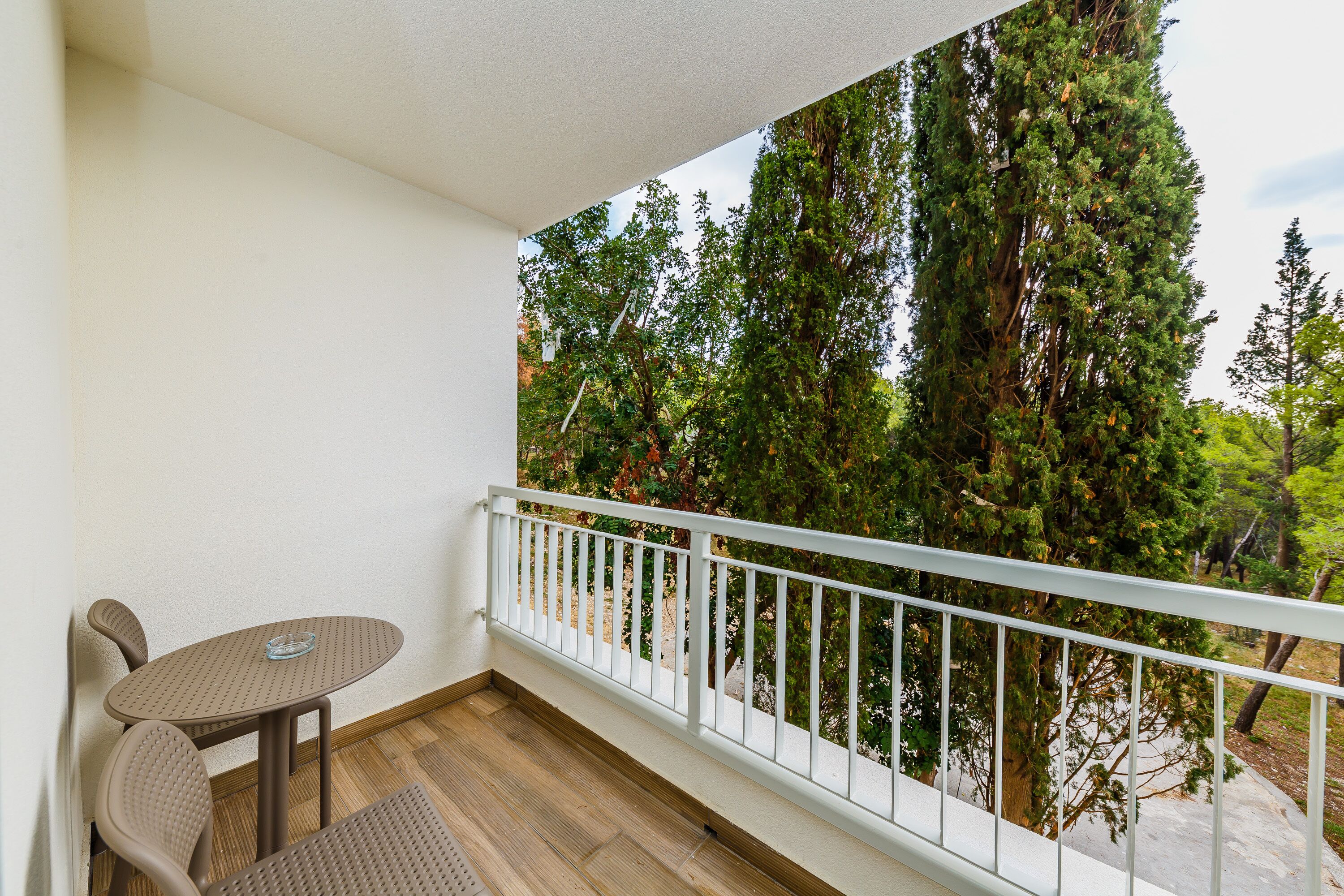 A balcony with a table and chairs, white railing, and a view of tall trees.
