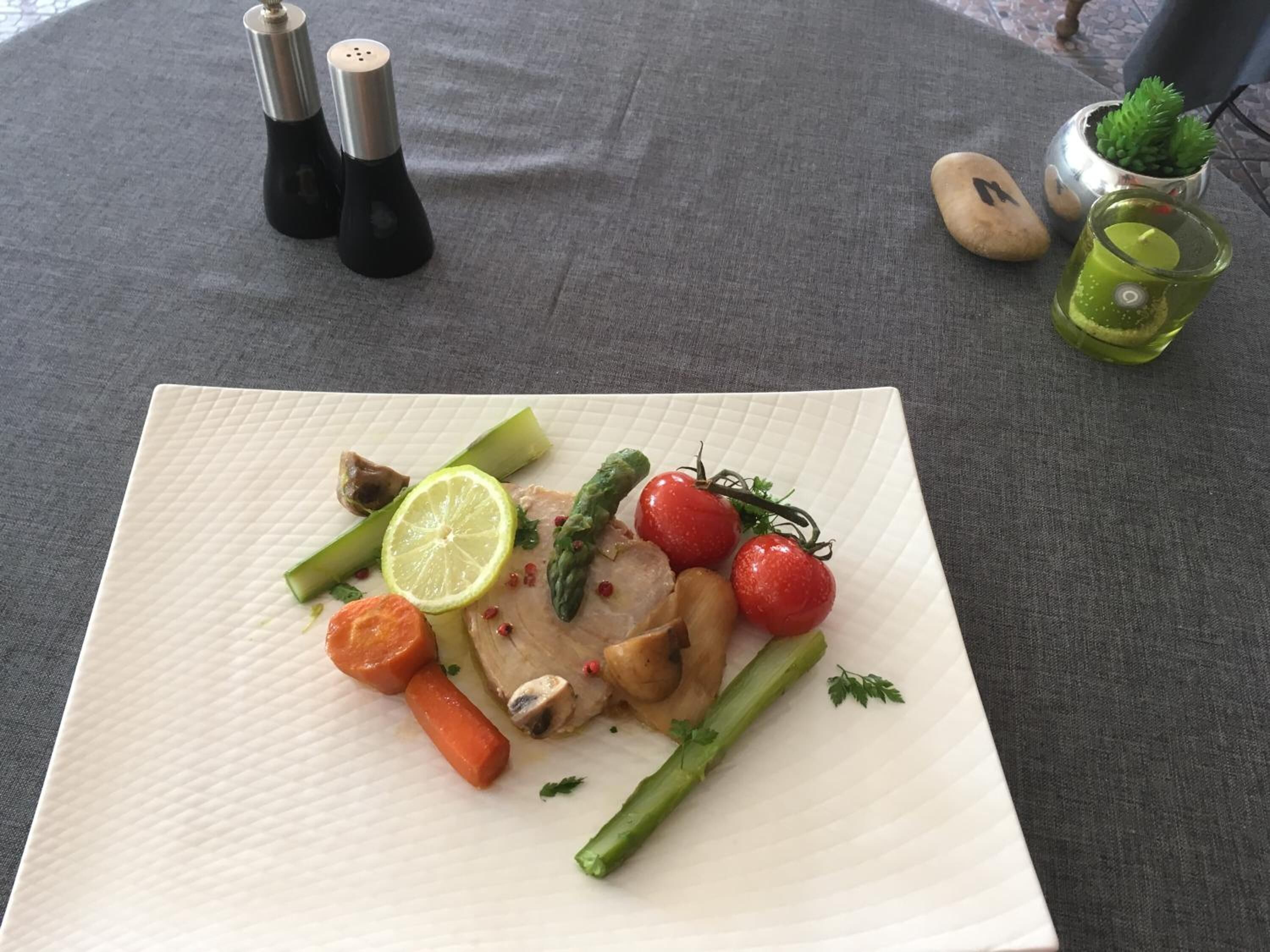 A white plate with a meal consisting of chicken, asparagus, cherry tomatoes, a carrot, and a slice of lime on a textured tablecloth.