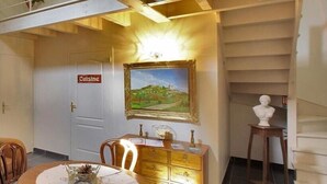 A dining area with a wooden table, chairs, a painting, a bust on a pedestal, and a staircase leading to an upper level.